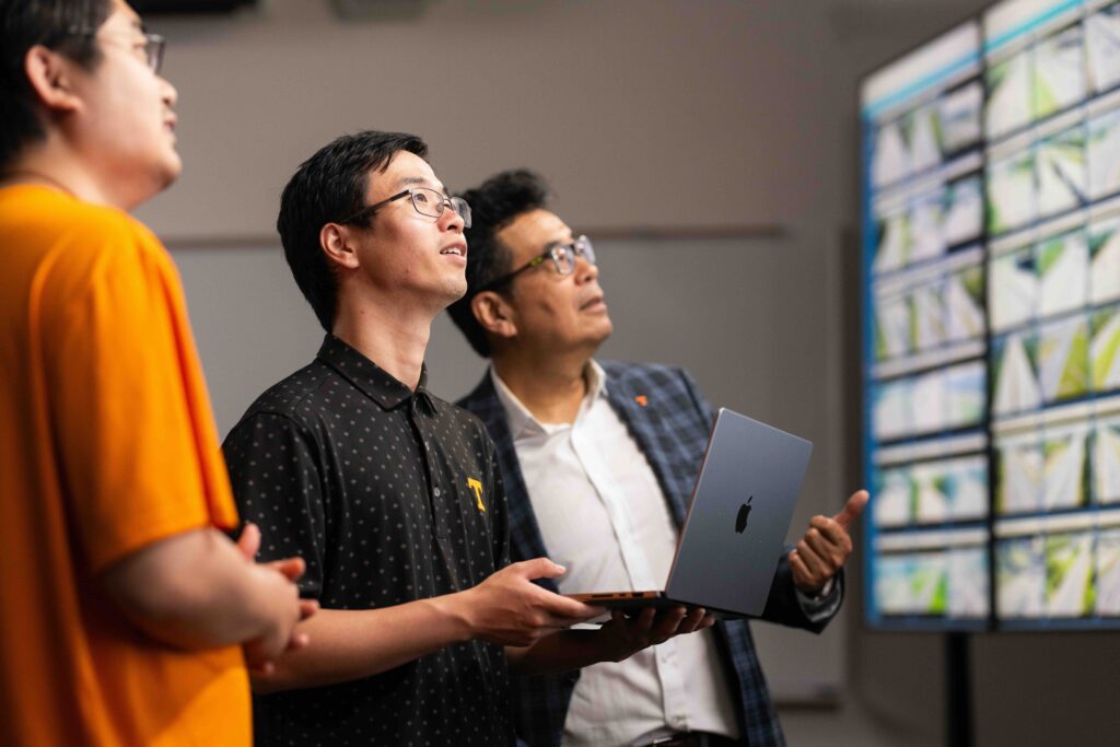 Professor Lee Han and two students in front of a bank of monitors, explaining the application of his research involving crowdsourced traffic and road condition data