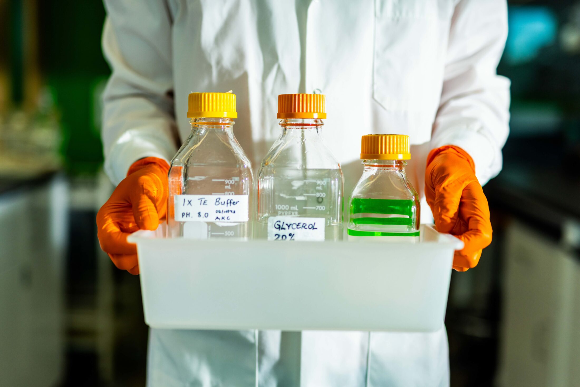 A detail of Alhagie K. Cham, Post Doc, hands as he carries bottles while wearing orange gloves in the Pathogen and Pest Lab inside the Agriculture and Natural Resources Building.