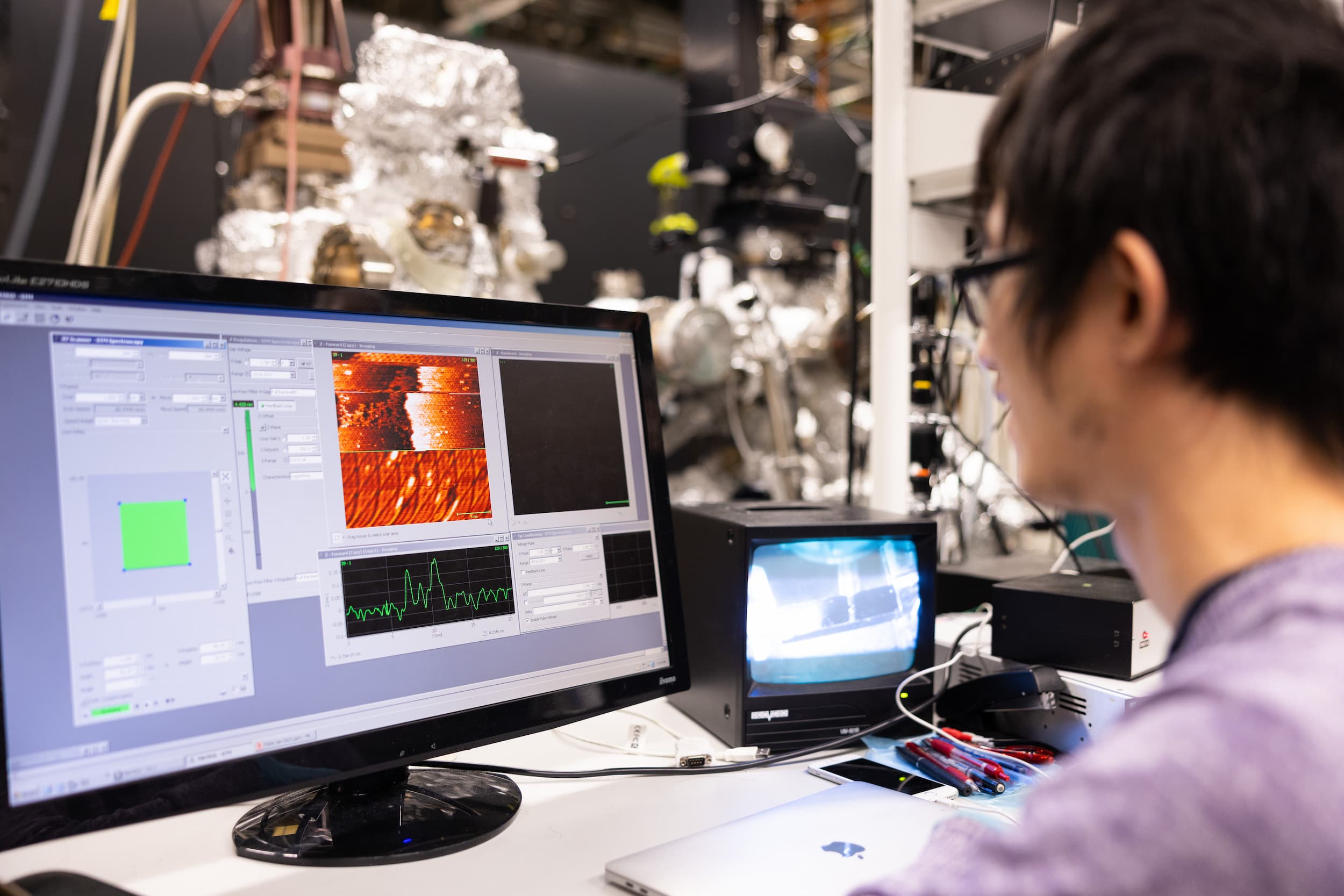 A male researcher wearing dark-rimmed glasses looks at a computer monitor as lab equipment operates.