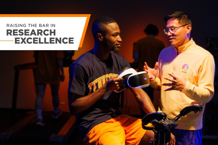 Professor and department head Zan Gao talks with kinesiology PhD student John Oginni simulating an experiment using a virtual reality bicycle exercise. Overlaid is a graphic with the text "Raising the bar in Research Excellence."