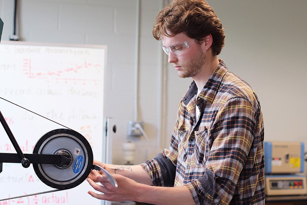A man in a flannel shirt wearing safety glasses grinds a blade at a belt sander in a manufacturing facility.
