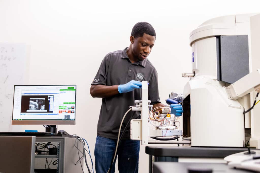 A black man works with a nano scale mechanical testing system integrated in a Scanning Electron Microscope inside an IAMM lab.