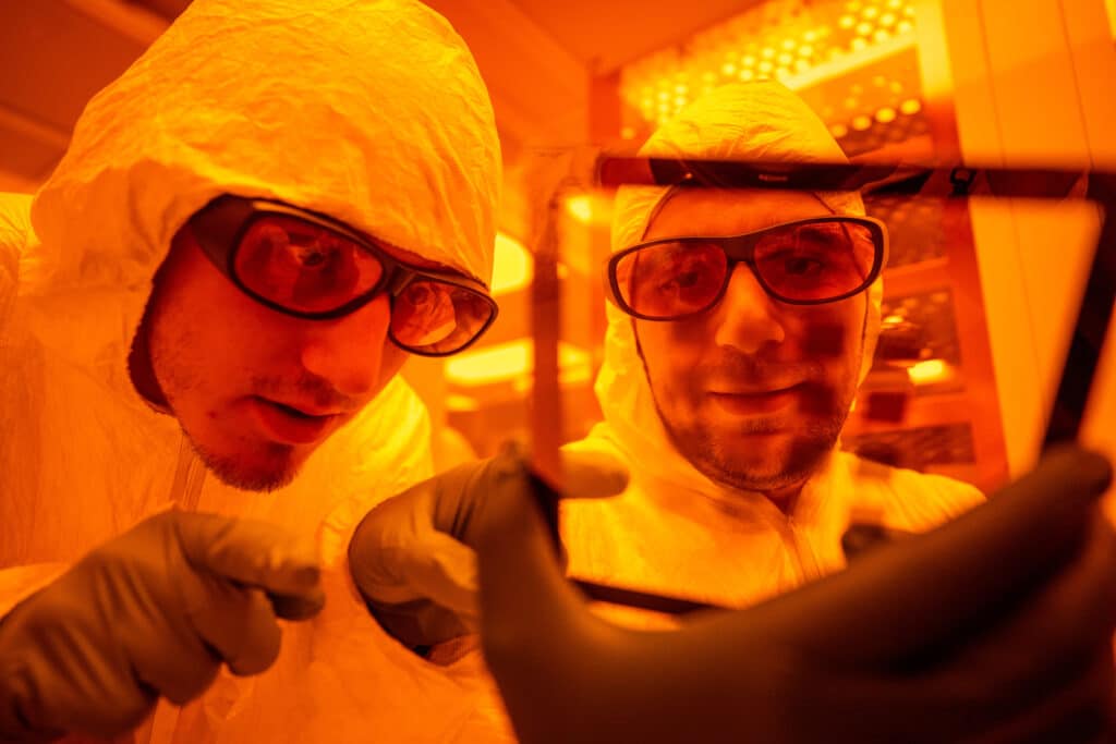 Two men in safety glasses, black gloves, and full, white protective body suits inspect a plate of transparent material in a lab. The image is tinted orange. 
