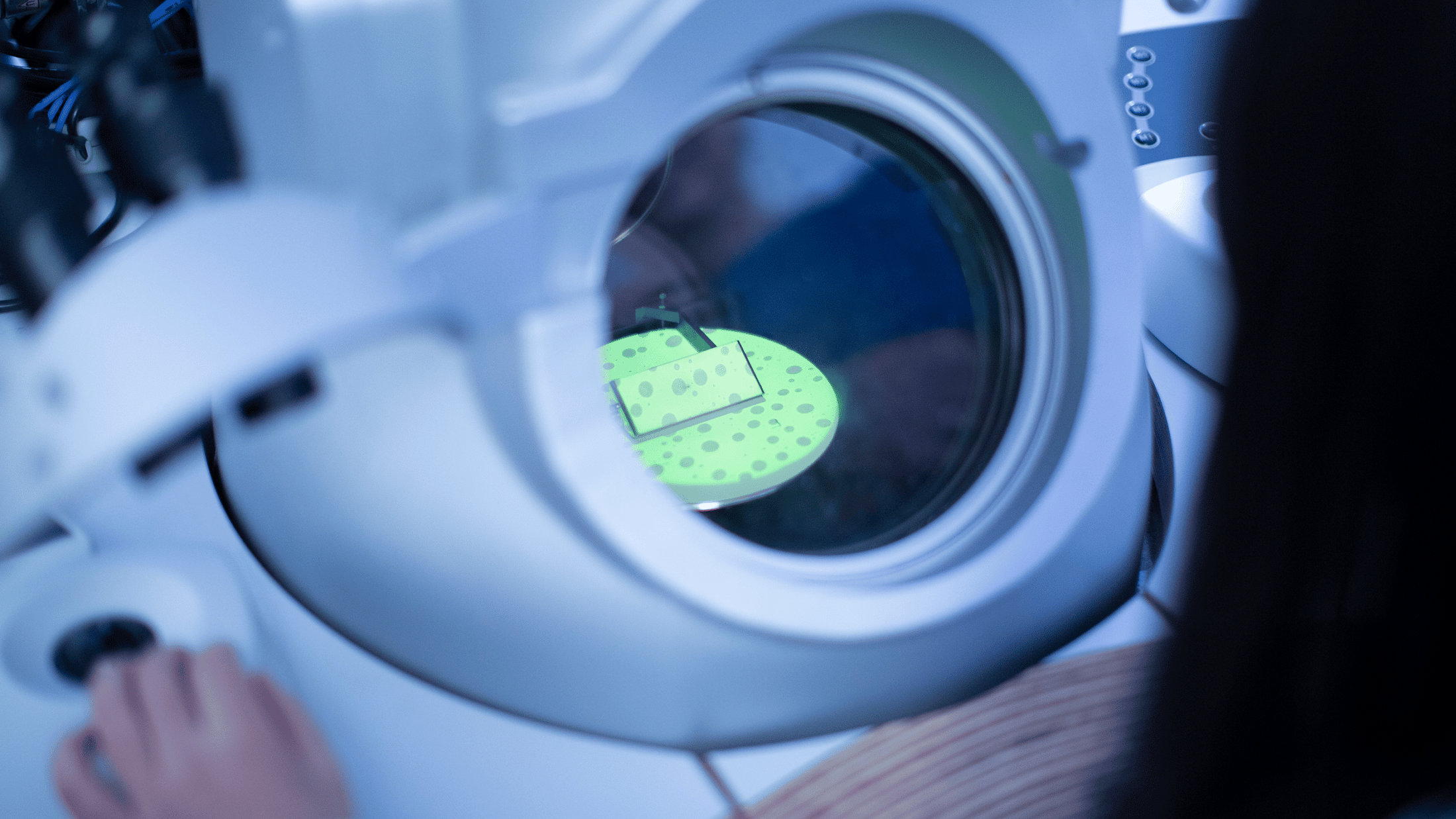 Close up of an electron microscope in use, a white machine with a circular view window looking in to a plate holding a specimen in a green light.