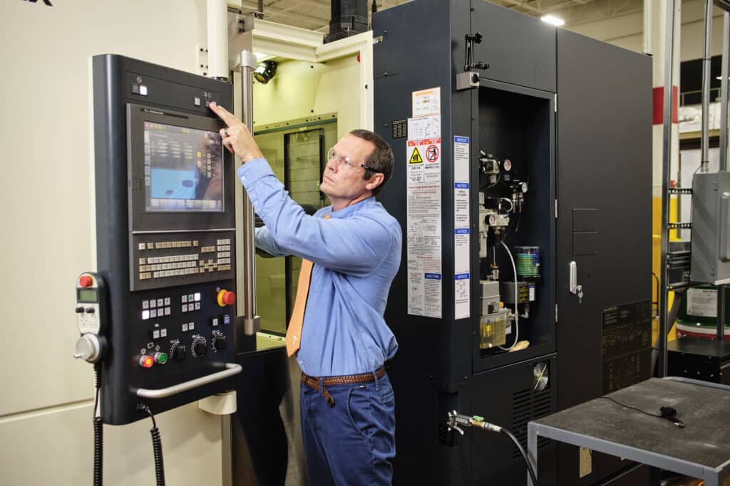 a white male professor in blue shirt and orange tie operates the buttons of a CNC milling machine in a lab filled with machinery. 