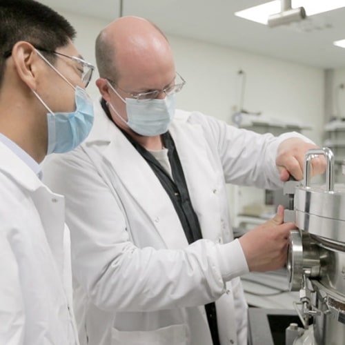 Two researchers wearing white lab coats and blue face masks work in a lab at the Institute for Advanced Materials and Manufacturing.