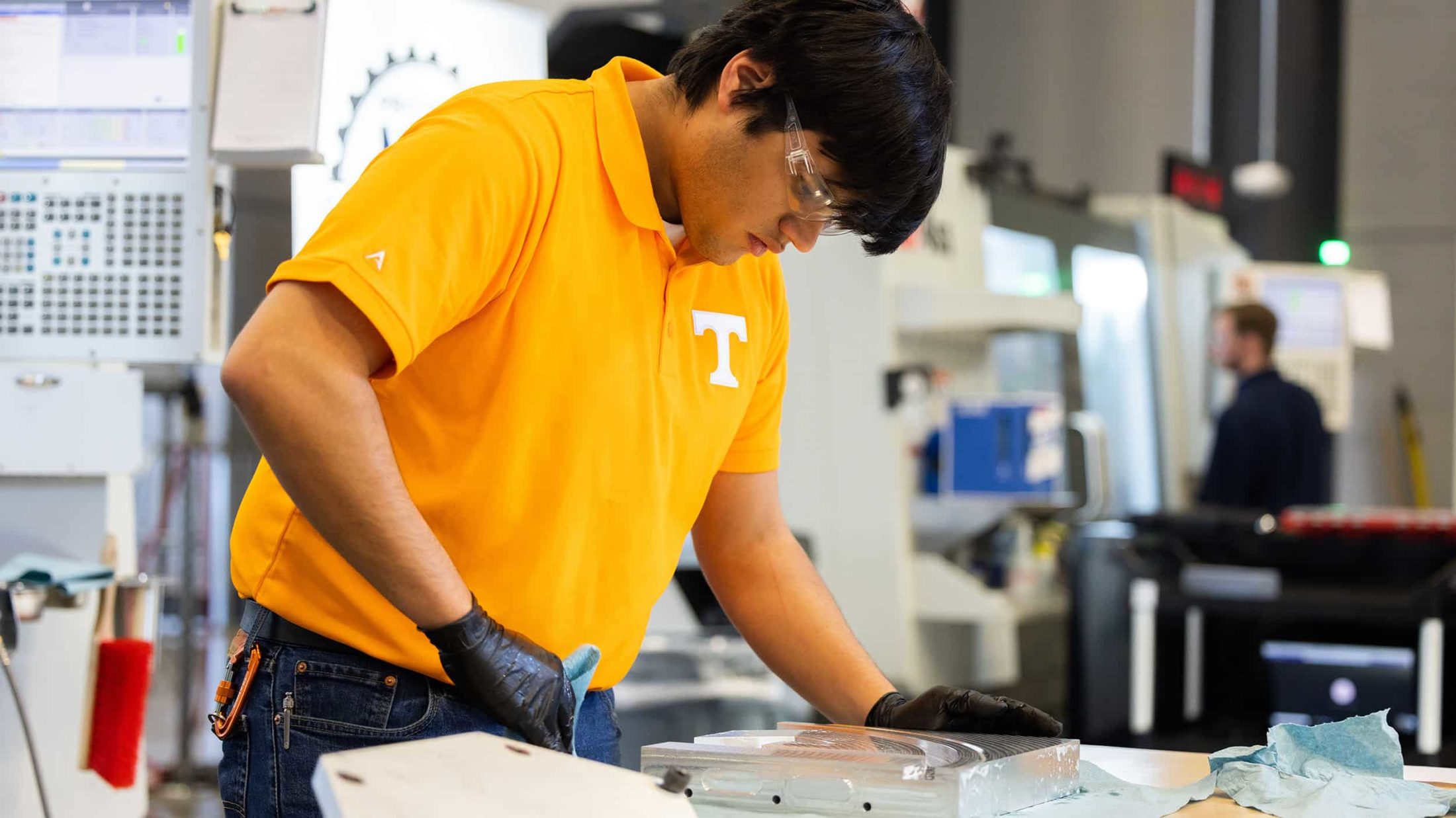 a student in an orange shirt polishes a shiny silver piece of metal on a table with large milling machines in the backround.