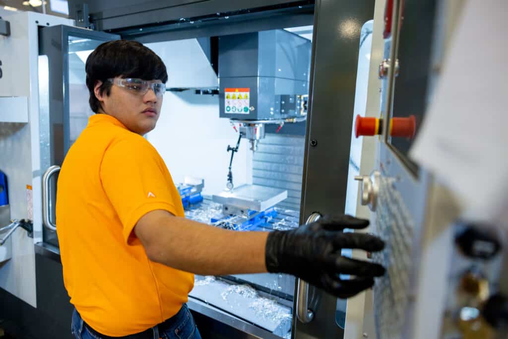 A male student in an orange polo shirt operates the buttons of a large HASS milling machine. Inside the machine sits a silver aluminum plate 