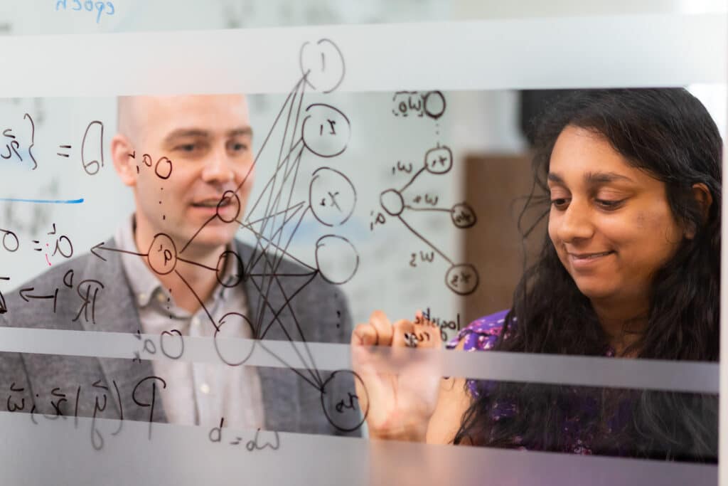 Two people, a white man and an Indian woman, collaborate behind a glass board, writing physics equations and diagrams on the glass. 