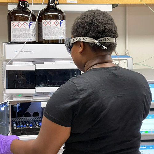 A Black woman wearing protective goggles operates lab equipment.
