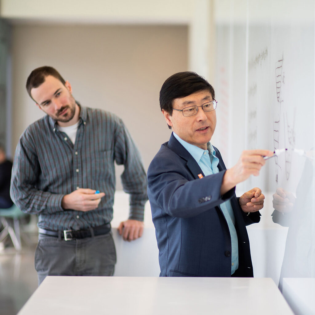 Bin Hu writes on a whiteboard at IAMM headquarters while a student watches.