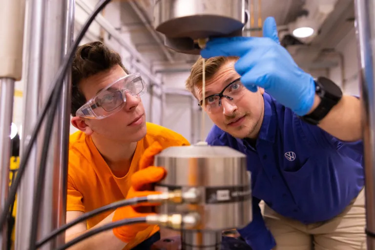 Two men wearing safety glasses work on glass fiber tensile equipment.