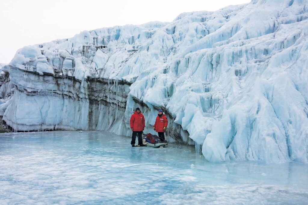 Two researchers wearing red winter coats stand in front of a glacier.