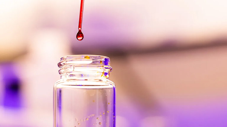 Close look at a pipette dropping a red liquid into a glass tube.