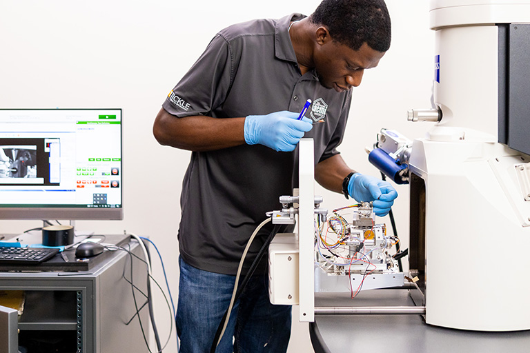 A researcher wearing a gray IACMI polo and gloves works on a scanning electron microscope.