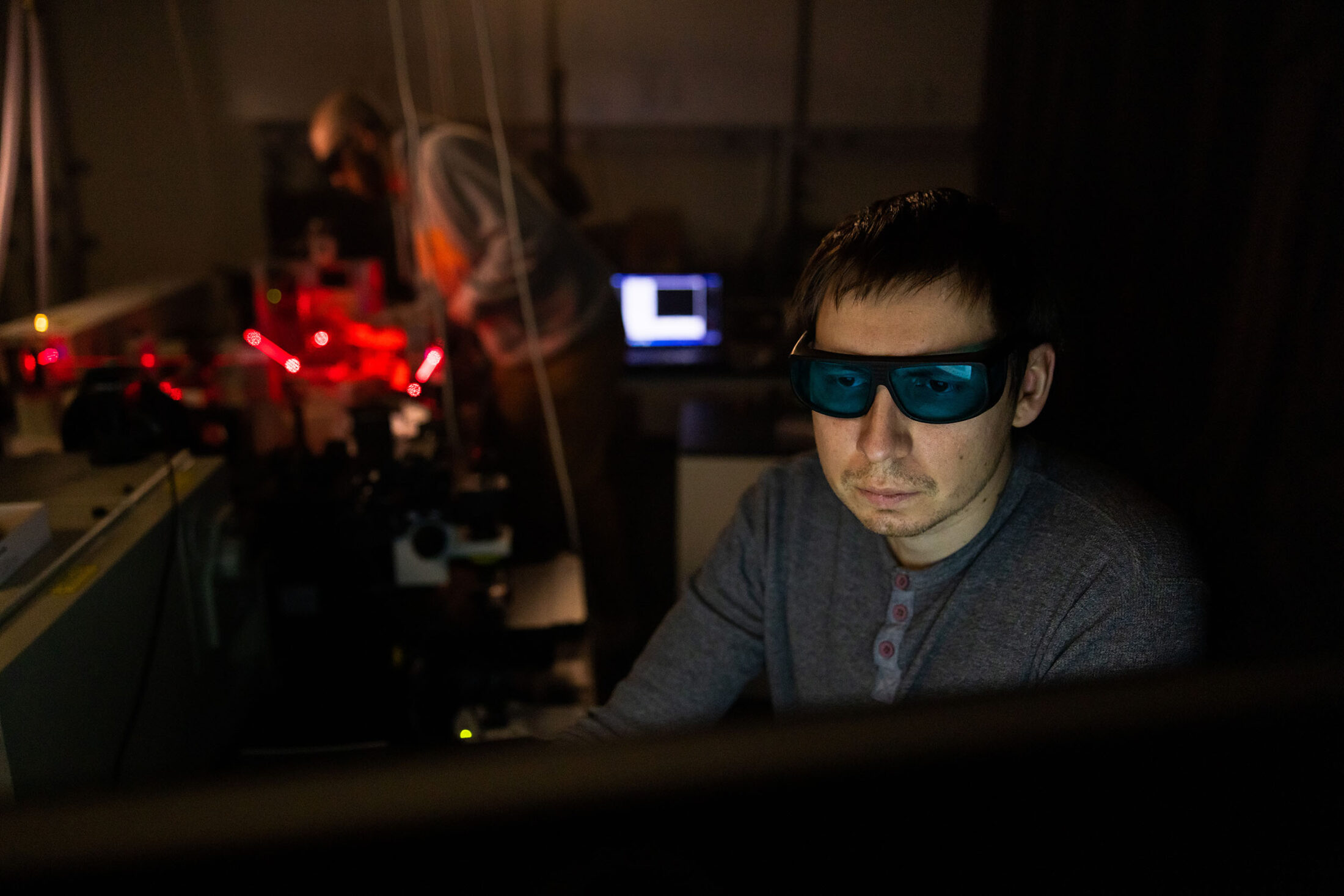 A student uses a computer as another student makes adjustments to a laser beam during an experiment.