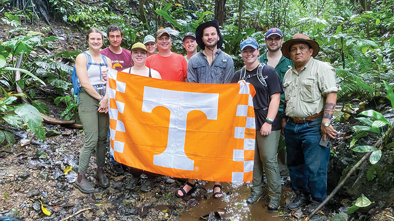 A group of students stand behind an orange flag with a white Power T on it in a wooded area in Panama.