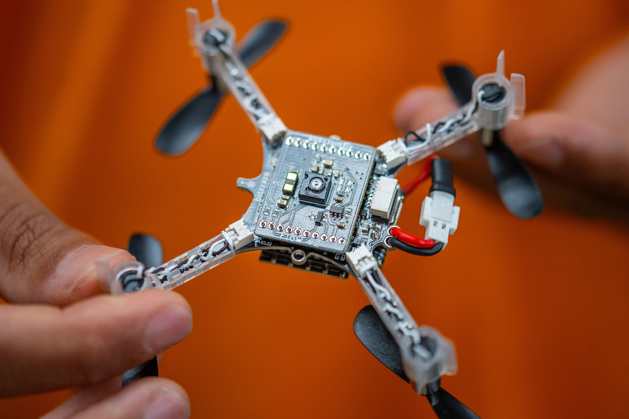 Two hands hold a nano drone with an orange shirt in the background.