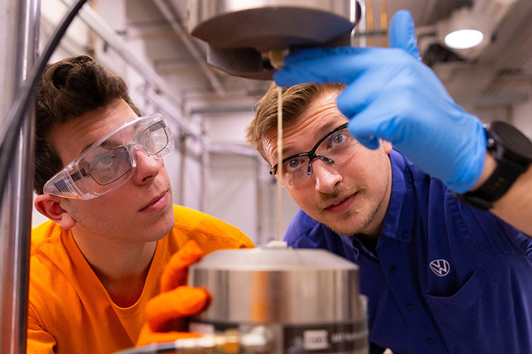 William Henken, a Volkswagen Doctoral Fellow, wearing a VW shirt, with a student about tensile testing of glass fiber reinforced polymeric (GFRP) composites in a lab inside the Science & Engineering Building at the University of Tennessee.