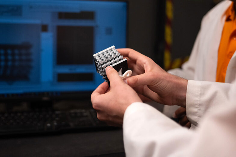 Two hands hold a 3D-printed materials sample in front of a monitor displaying the same sample during testing.