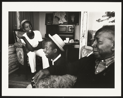 A black-and-white photograph of James Baldwin, wearing a white hat, sitting on the floor at center. On the right, Beauford Delaney sits in a chair. On the left, the daughter of Bertice Reading wears light colors. Source: National Museum of African American History and Culture