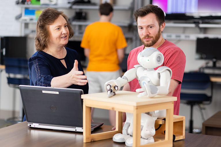 While talking to a student, Lynne Parker gestures at a robot sitting at a small desk on a table.