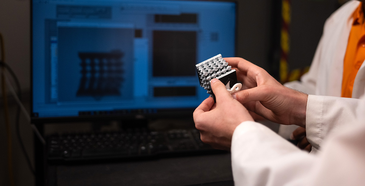 A graduate student holds a 3D printed sample in front of a monitor displaying the same sample during testing.