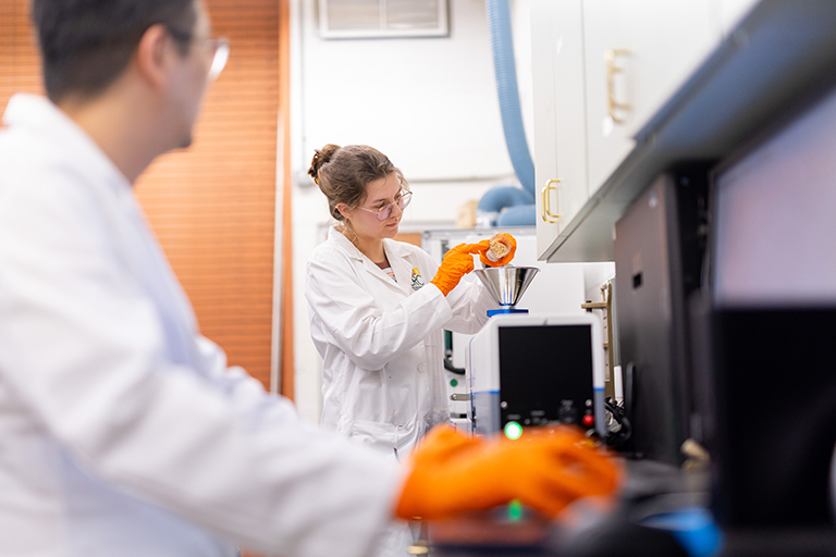 A student pours a biomass sample into a particle-sized 3D image analyzer as a PhD student reviews the results.