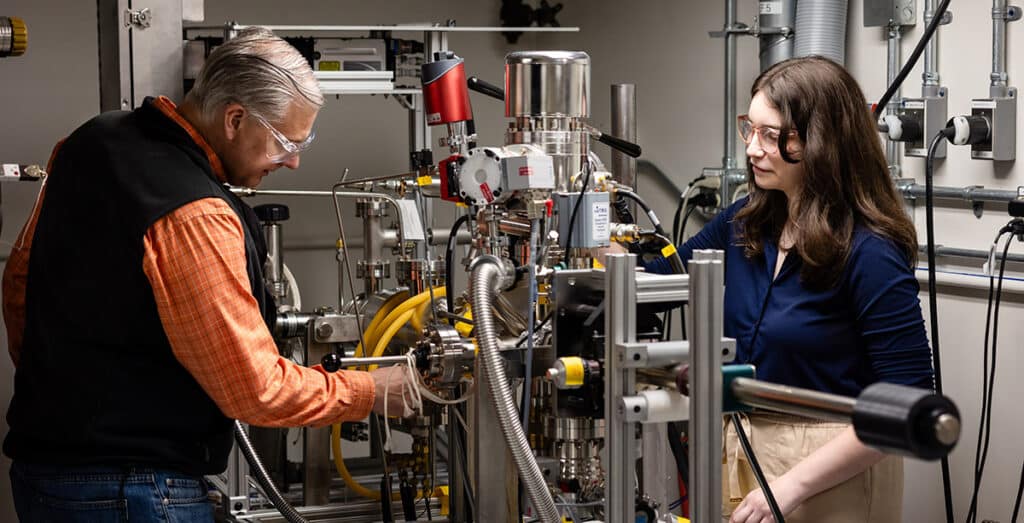 Two researchers in safety glasses adjusting complex metal nuclear equipment in a science laboratory.