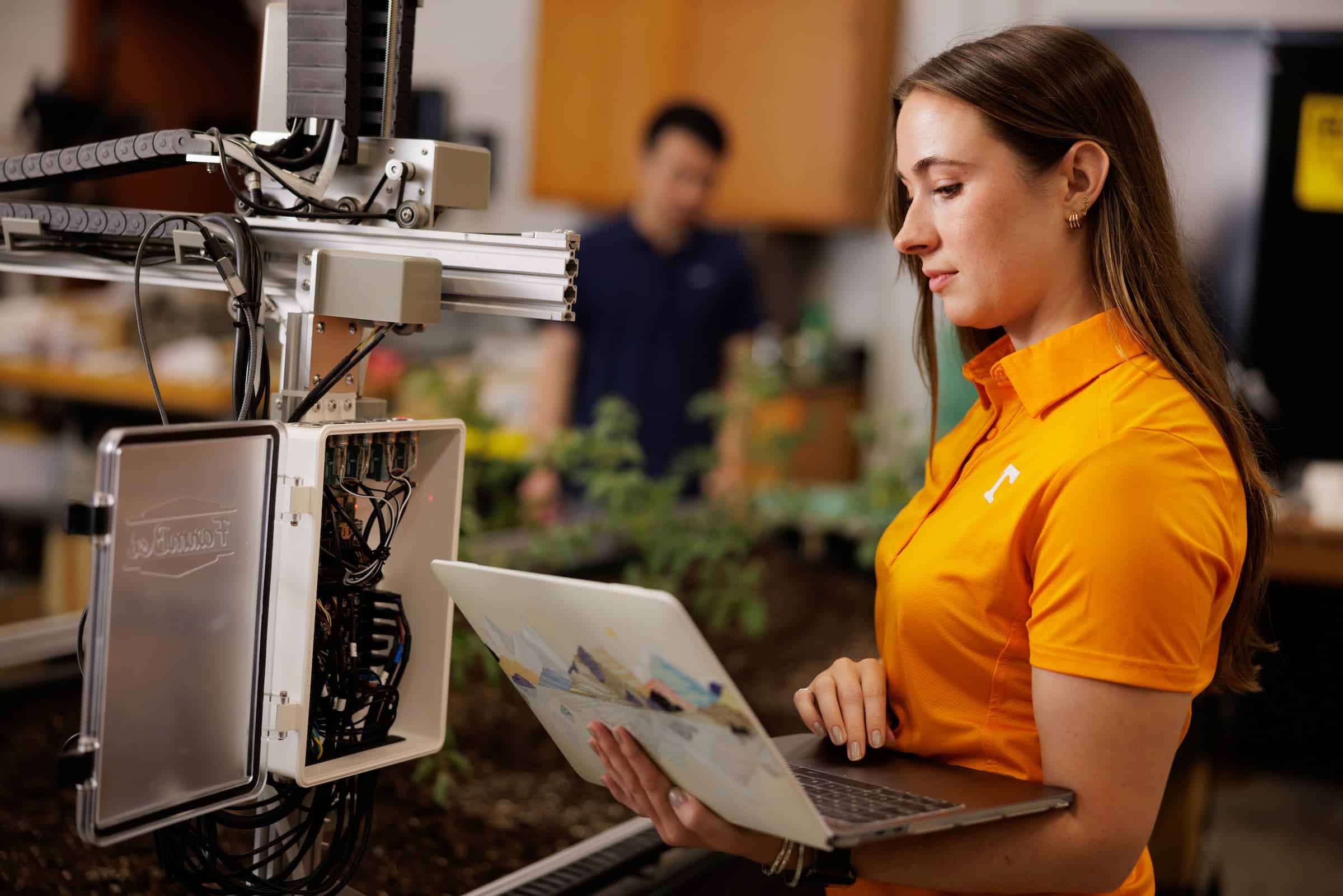A researcher in an orange shirt uses a laptop to monitor automated agricultural equipment in a lab.