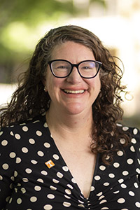 A smiling person with curly hair and glasses wears a black polka-dot top outdoors, with trees and soft light in the background.