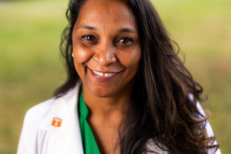 A smiling person with long dark hair wears a white coat with an orange "T" logo over a green shirt, standing outdoors against a blurred green background.