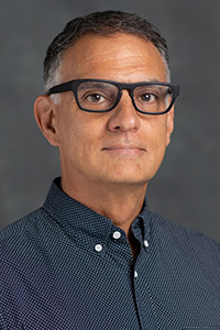 A person with short hair and glasses wears a dark patterned button-up shirt against a textured gray background.